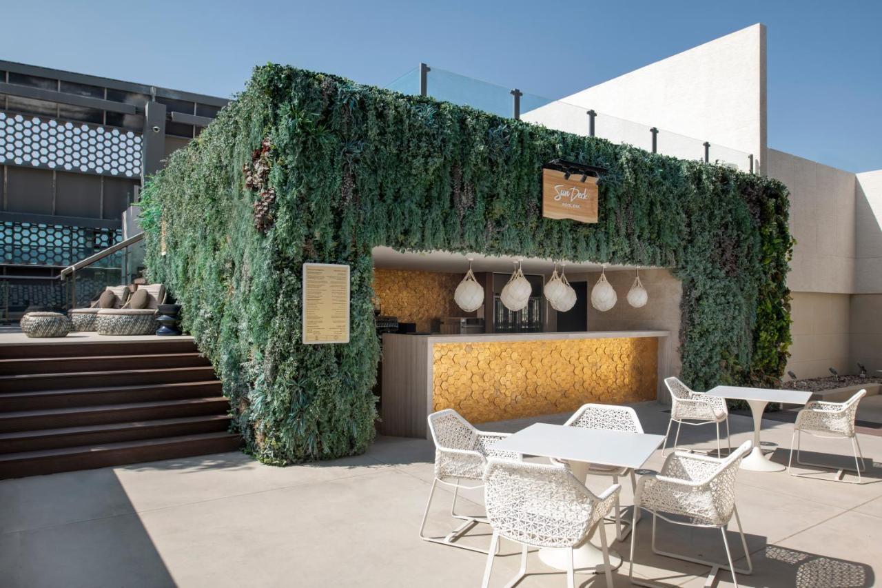 Steigenberger Hotel Doha Kültér fotó The photo shows an outdoor bar area surrounded by lush greenery. The bar has a modern design, featuring a wooden counter with decorative pendant lights hanging above. There are several white outdoor tables and chairs arranged in front of the bar, whi